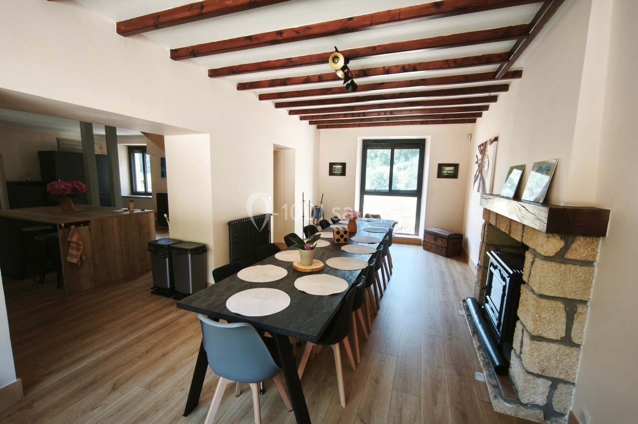 Salle à manger lumineuse avec une grande table en bois, des chaises modernes, une cheminée en pierre et des poutres…