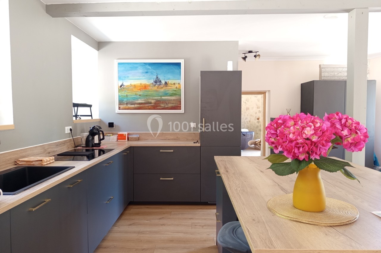 Cuisine moderne avec meubles gris, plan de travail en bois, tableau coloré au mur et vase de fleurs roses sur l'îlot.