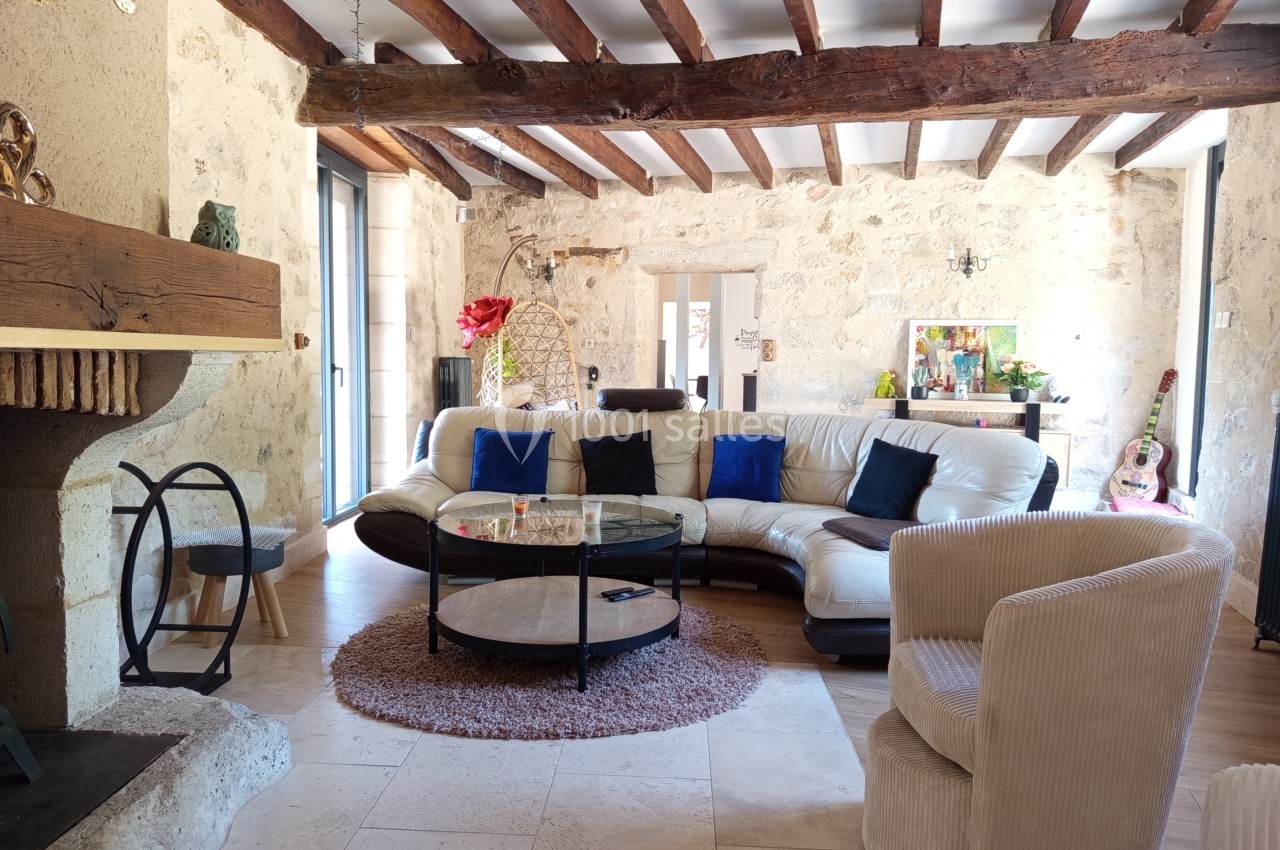 Salon lumineux avec murs en pierre, poutres apparentes, canapé en cuir blanc, cheminée et décoration chaleureuse.