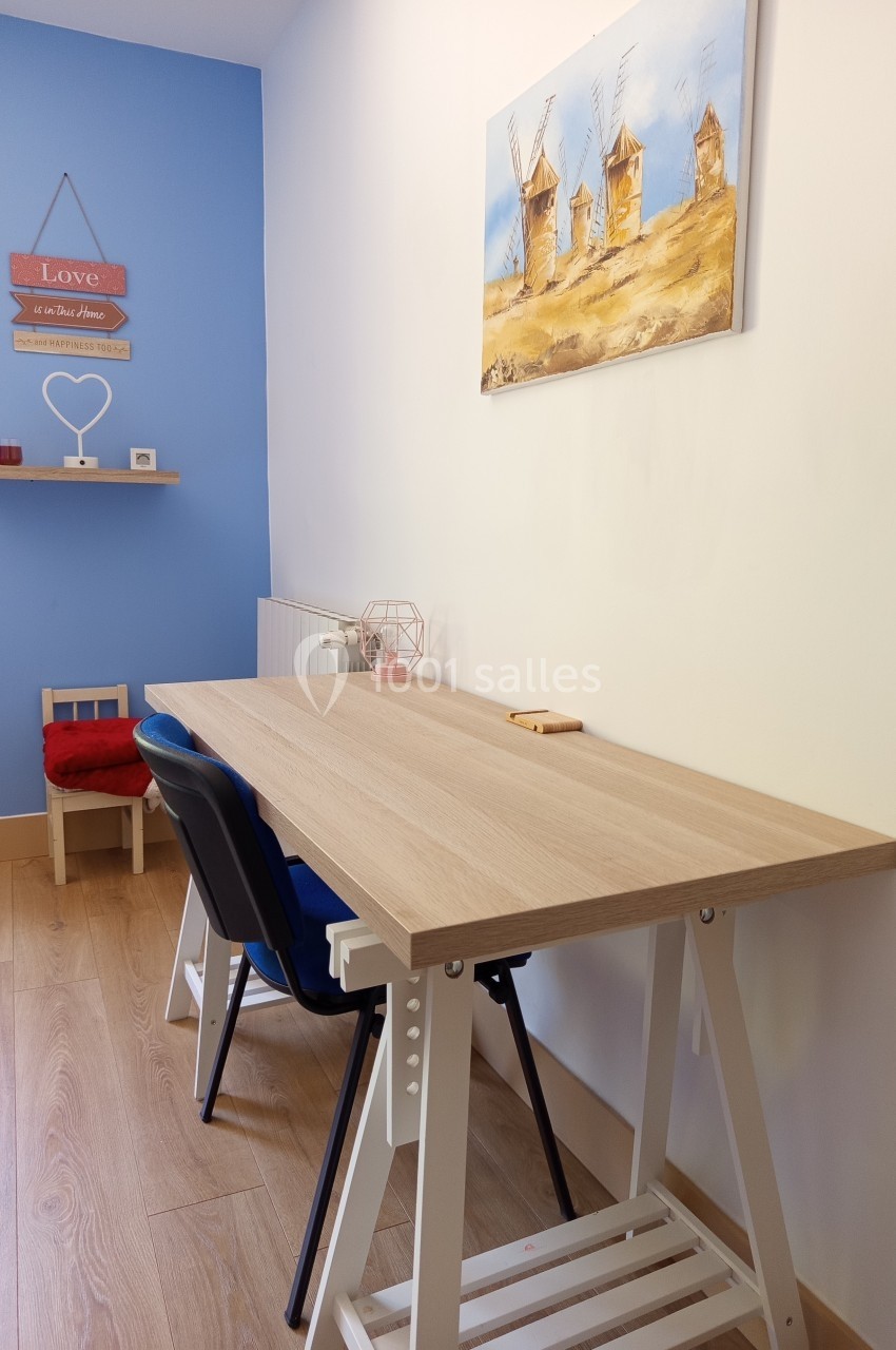 Bureau en bois clair avec deux chaises, situé dans une pièce aux murs bleus et blancs, décorée de cadres et d'étagères.