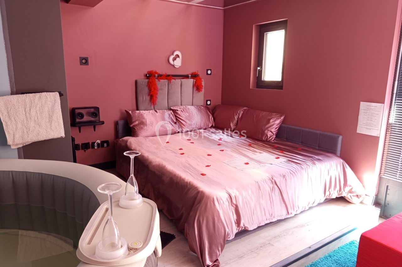 Chambre avec lit double décoré de pétales rouges, bain à remous au premier plan et lumière naturelle entrant par une fenêtre.