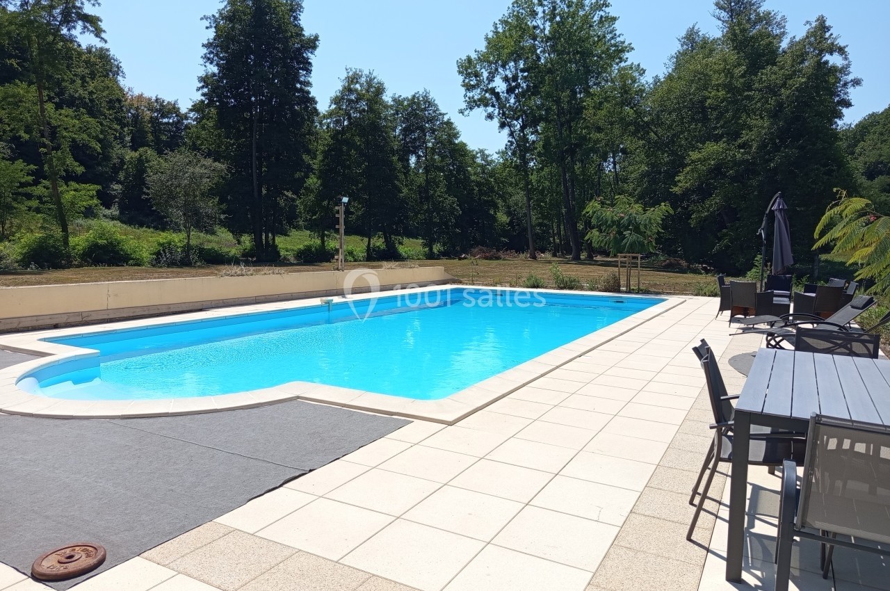 Piscine extérieure entourée de dalles claires, avec des chaises et une table à proximité, bordée par des arbres.
