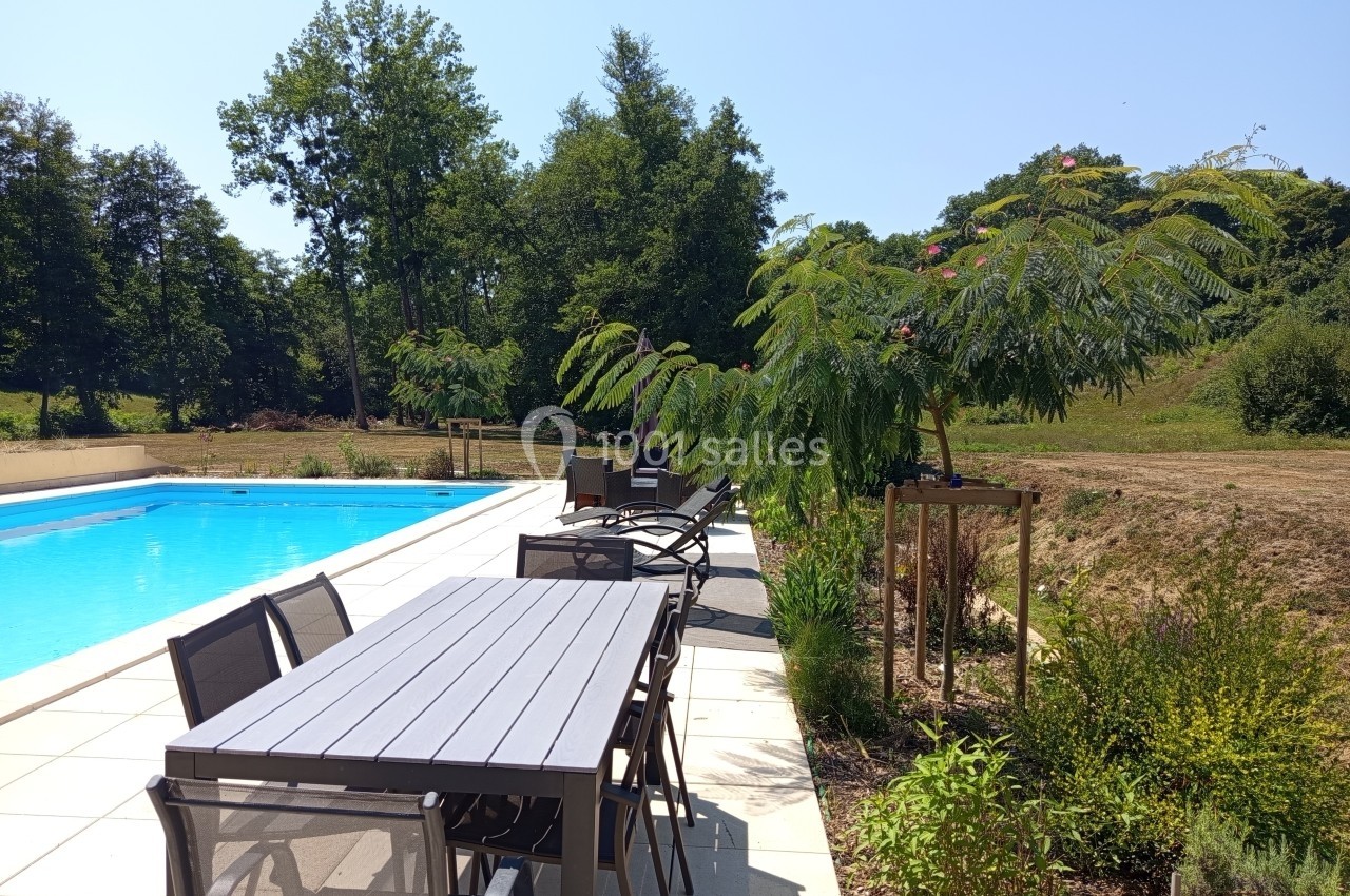 Terrasse avec table et chaises en métal, bordant une piscine entourée de végétation et d'arbres sous un ciel dégagé.