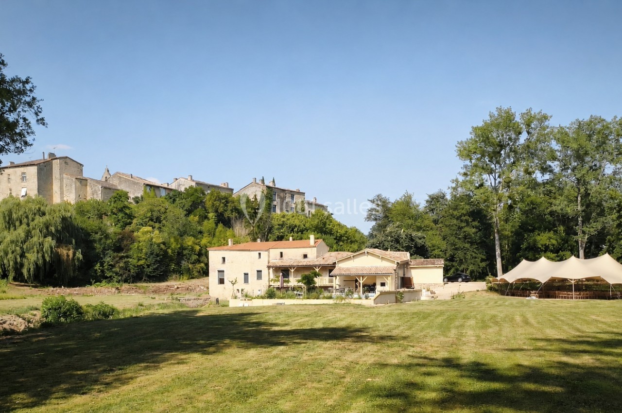 Vue d'un domaine avec bâtiments en pierre, grande pelouse, tente blanche et village perché en arrière-plan.