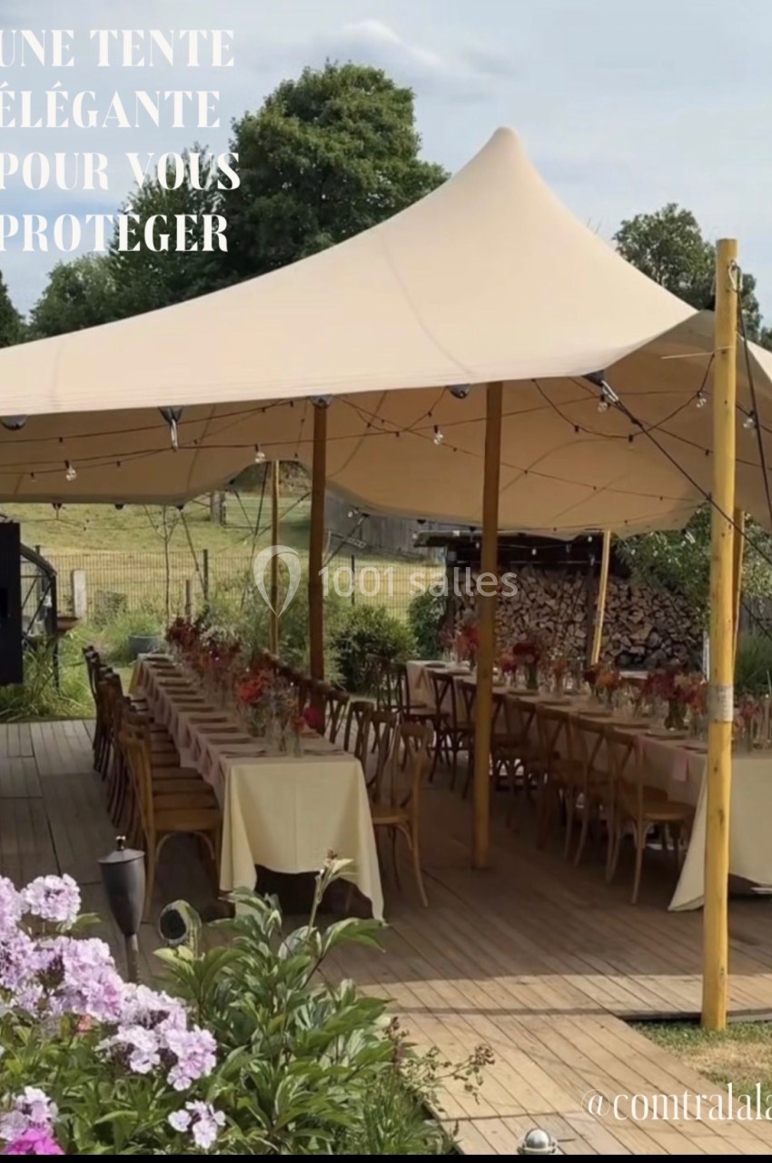 Tente beige installée en extérieur, couvrant des tables et chaises en bois, entourée de verdure et de fleurs.