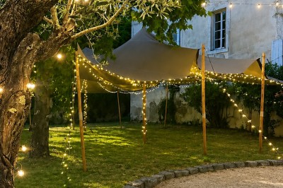 Tables décorées pour un dîner en plein air sous une tente éclairée de guirlandes lumineuses, près d'un bâtiment en pierre.