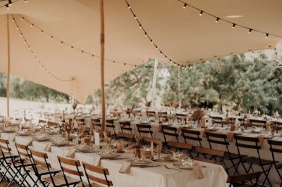 Tables décorées pour un dîner en plein air sous une tente éclairée de guirlandes lumineuses, près d'un bâtiment en pierre.
