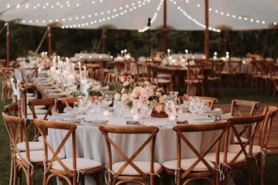 Tables décorées pour un dîner en plein air sous une tente éclairée de guirlandes lumineuses, près d'un bâtiment en pierre.