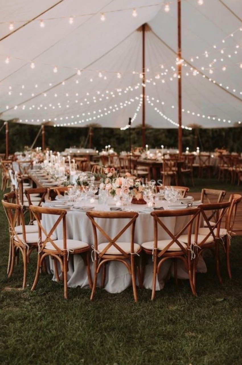 Tables rondes décorées avec des fleurs et des bougies sous une tente éclairée par des guirlandes lumineuses.