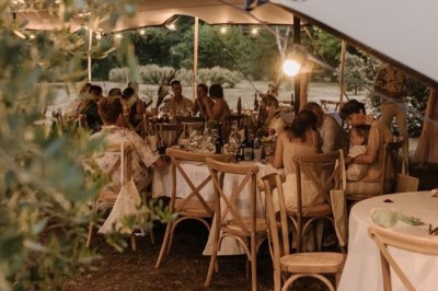 Tables décorées pour un dîner en plein air sous une tente éclairée de guirlandes lumineuses, près d'un bâtiment en pierre.