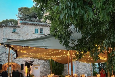 Tables décorées pour un dîner en plein air sous une tente éclairée de guirlandes lumineuses, près d'un bâtiment en pierre.