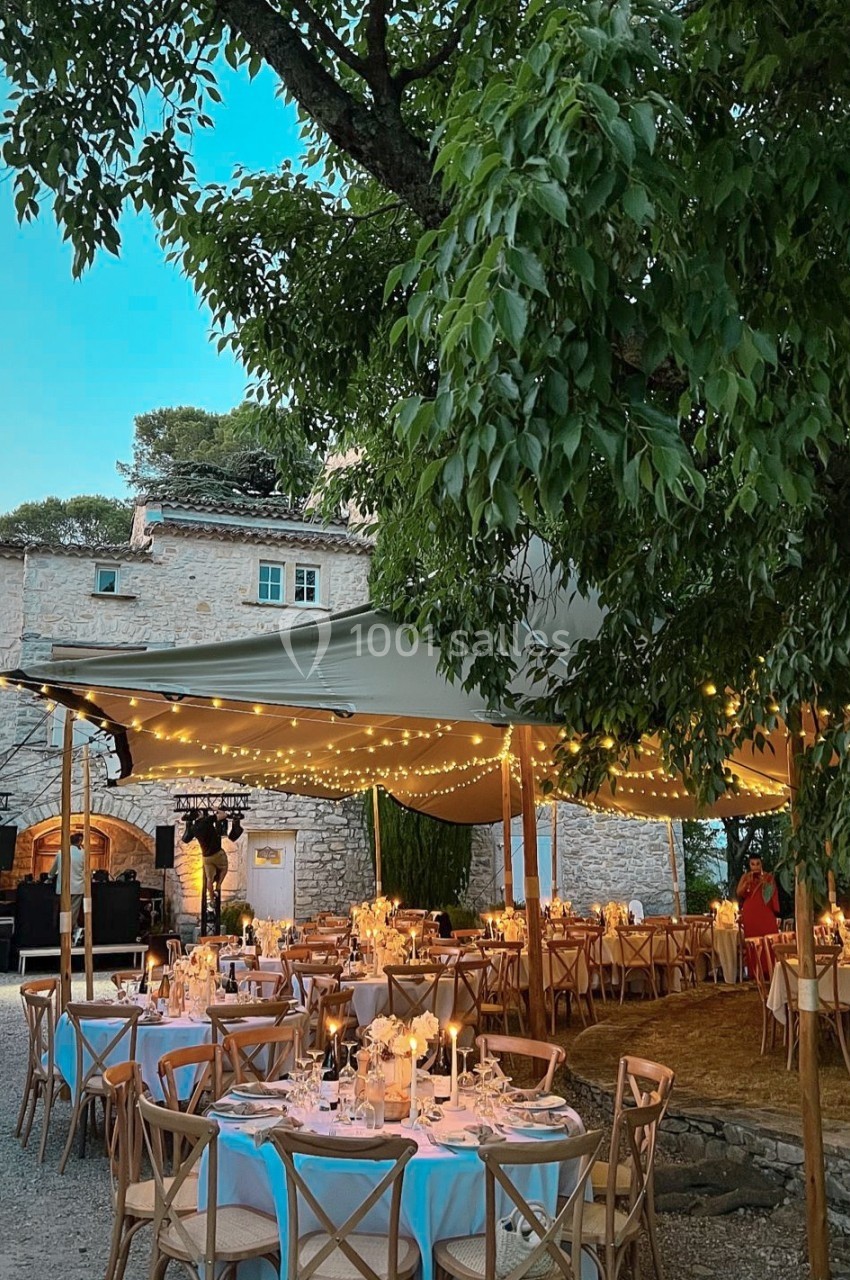 Tables décorées pour un dîner en plein air sous une tente éclairée de guirlandes lumineuses, près d'un bâtiment en pierre.