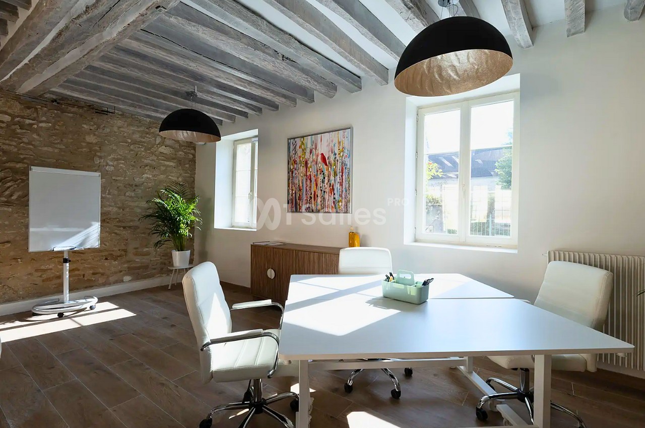 Salle de réunion lumineuse aux poutres apparentes et murs en pierre, avec une grande table blanche entourée de chaises de bureau blanches.