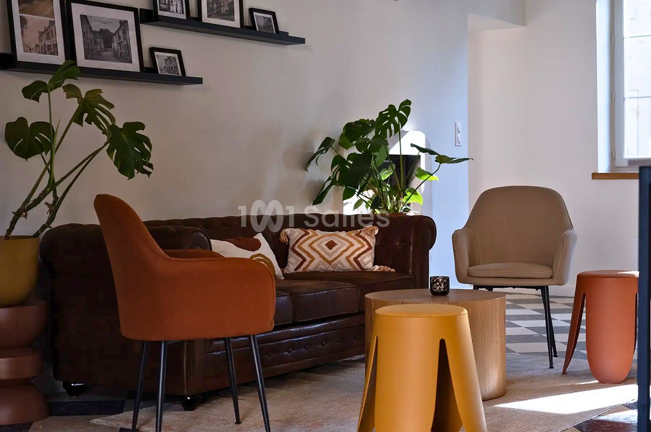 Salon lounge au sol carrelé en damier, avec un canapé Chesterfield en cuir brun, des fauteuils colorés et des tables basses en bois.