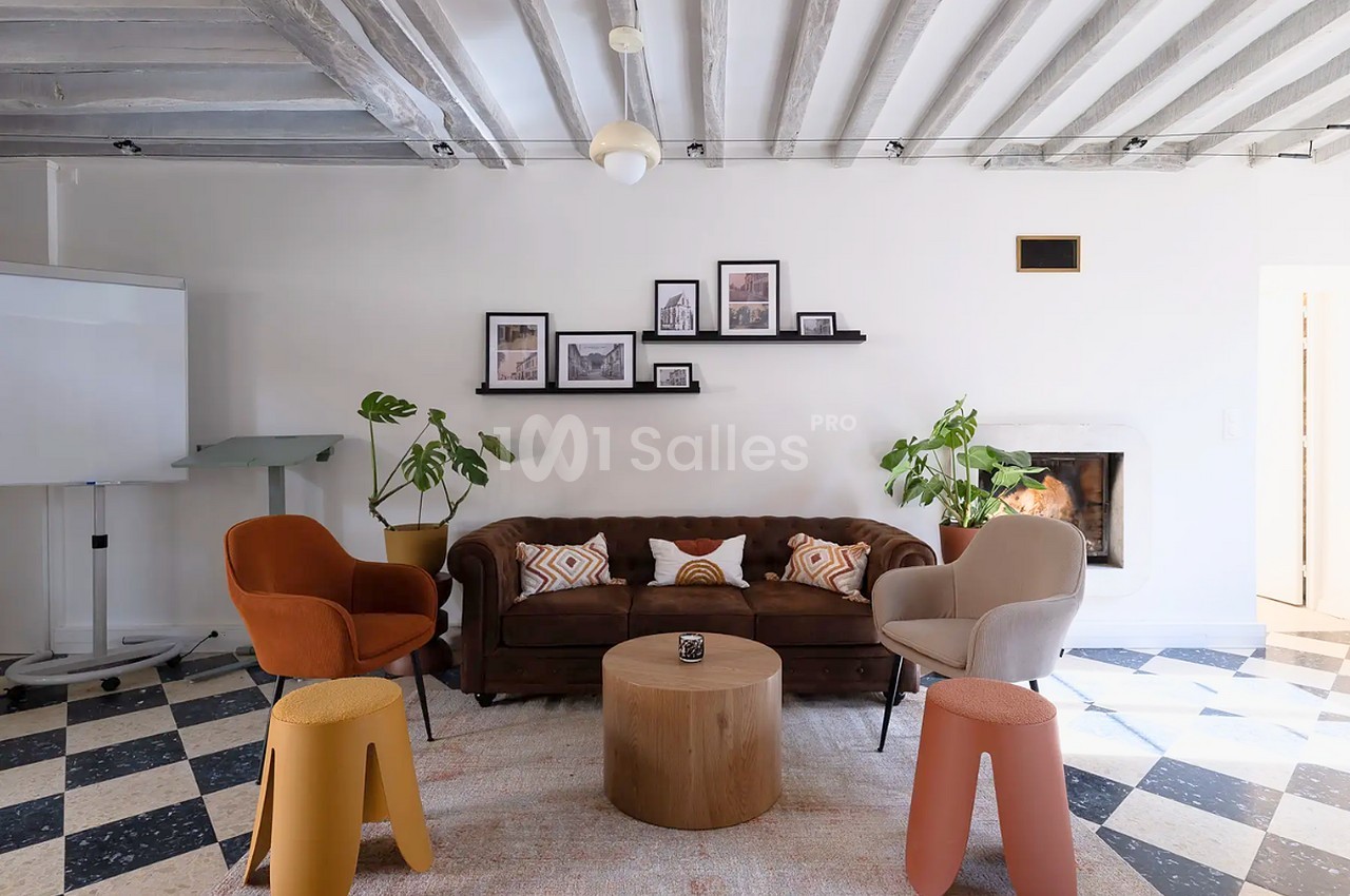 Salon aux poutres apparentes, avec canapé Chesterfield brun, fauteuils colorés, poufs et table basse en bois, près d'une cheminée et d'un tableau blanc.