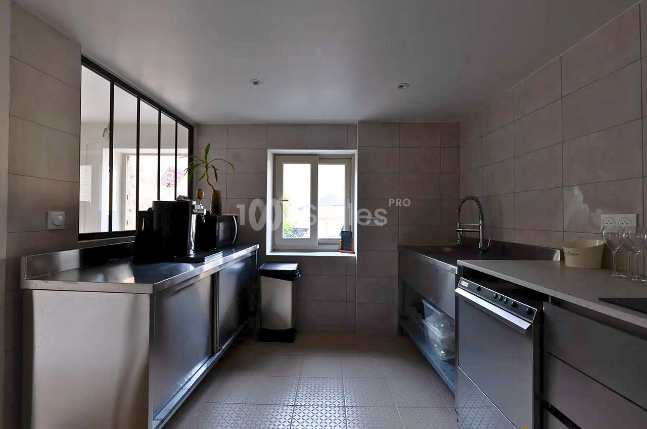 Cuisine professionnelle en inox à deux plans de travail parallèles, avec lave-vaisselle, carrelage mural beige et fenêtre au fond.