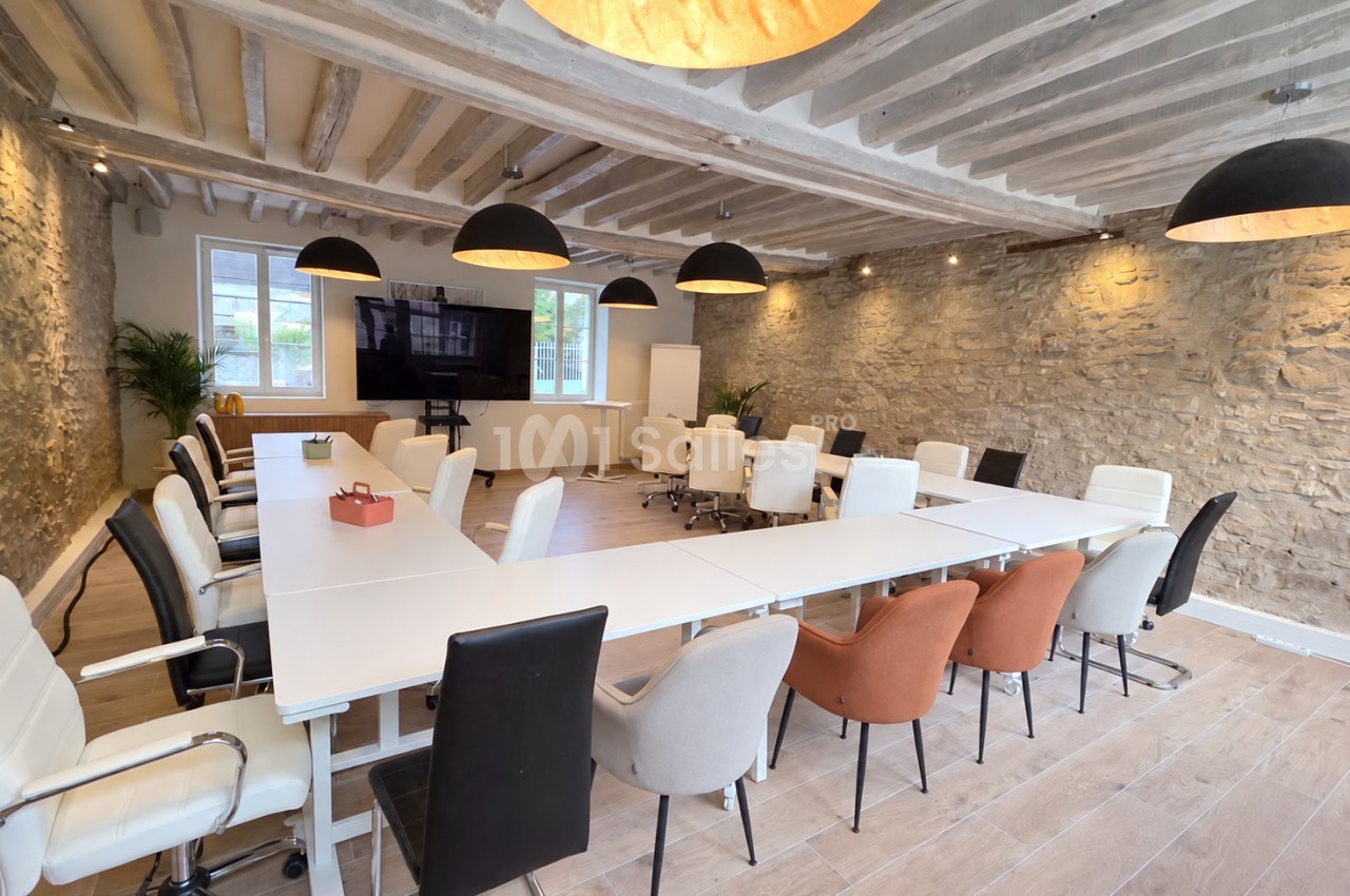 Salle de réunion aux poutres apparentes et murs en pierre, avec une grande table blanche en U entourée de chaises noires, blanches et terracotta.