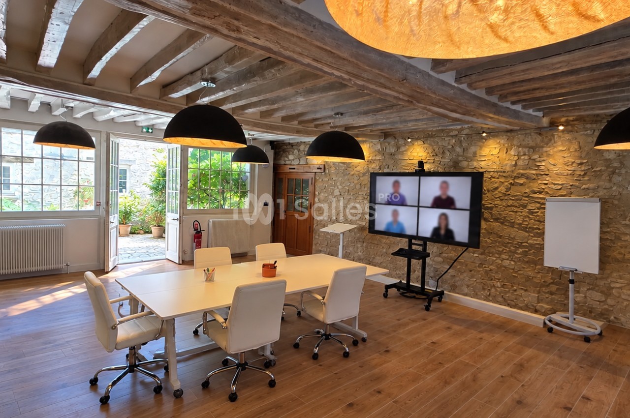 Salle de réunion aux poutres apparentes et mur de pierre, avec table blanche, chaises, écran de visioconférence et chevalet.