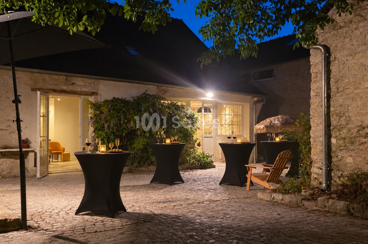 Cour pavée éclairée la nuit, avec quatre tables hautes habillées de noir, verres et bougies posés, et un bâtiment en pierre aux portes ouvertes.