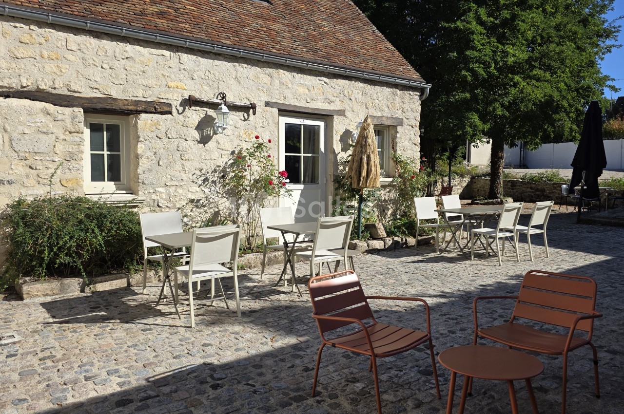 Terrasse pavée en extérieur avec tables et chaises blanches devant une façade en pierre apparente, et deux fauteuils et une petite table rouille au premier plan.