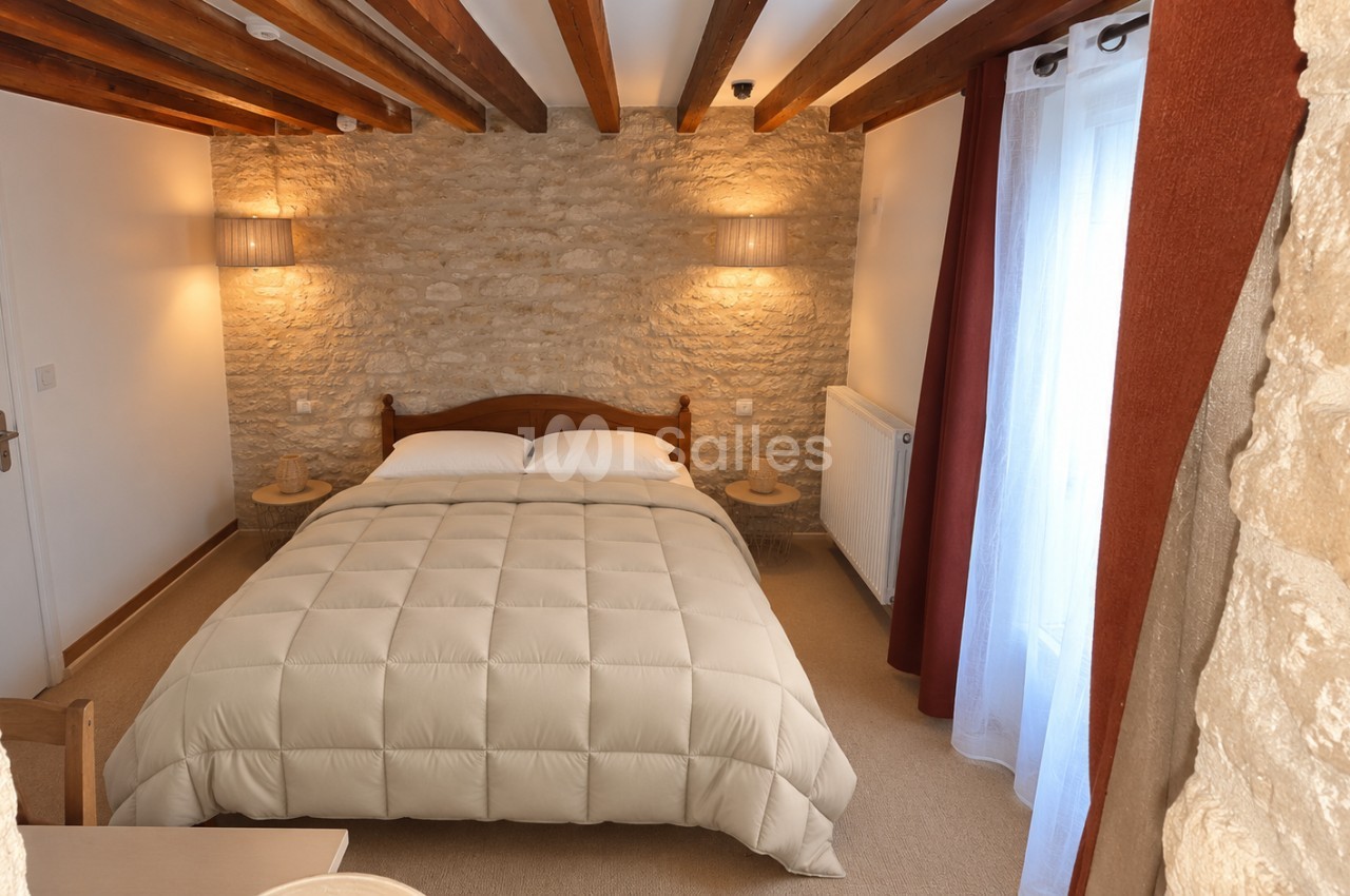 Chambre avec un grand lit recouvert d'une couette beige, mur en pierre apparente, poutres en bois au plafond et rideaux rouges.