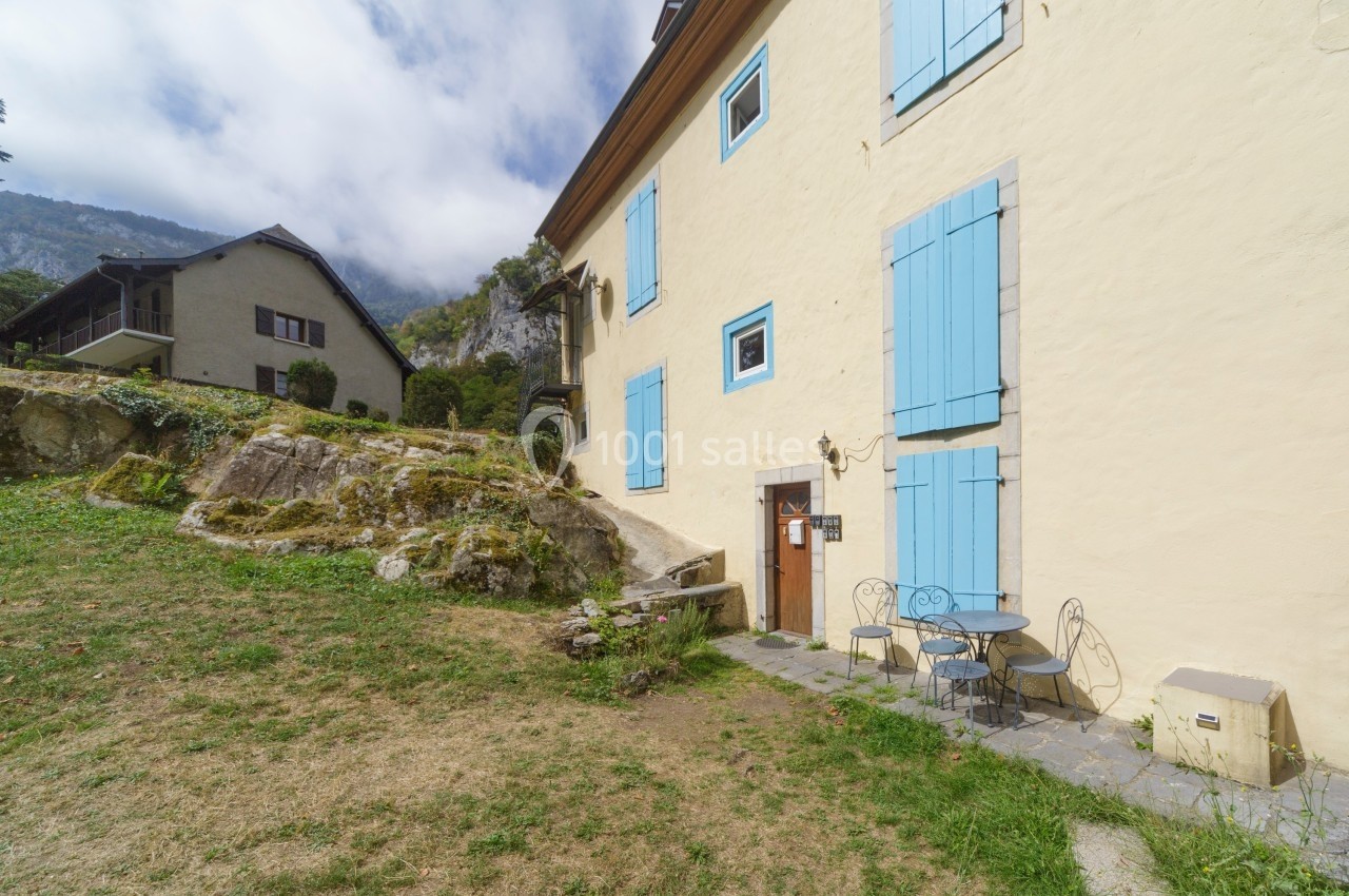 Façade d'une maison beige avec volets bleus, table et chaises en fer forgé sur une terrasse en pierre, jardin en pente.