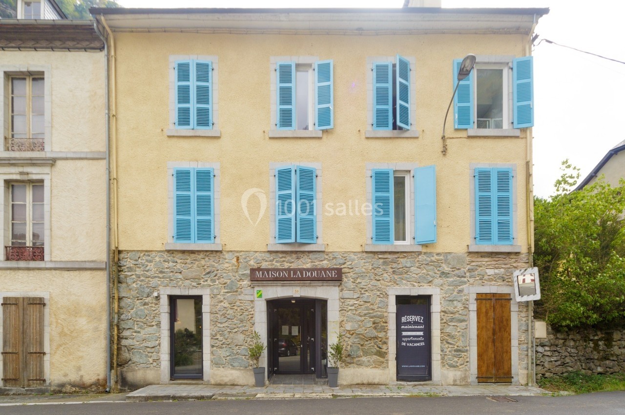 Façade d'une maison en pierre avec volets bleus, une enseigne ’Maison La Douane’ et une entrée principale vitrée.