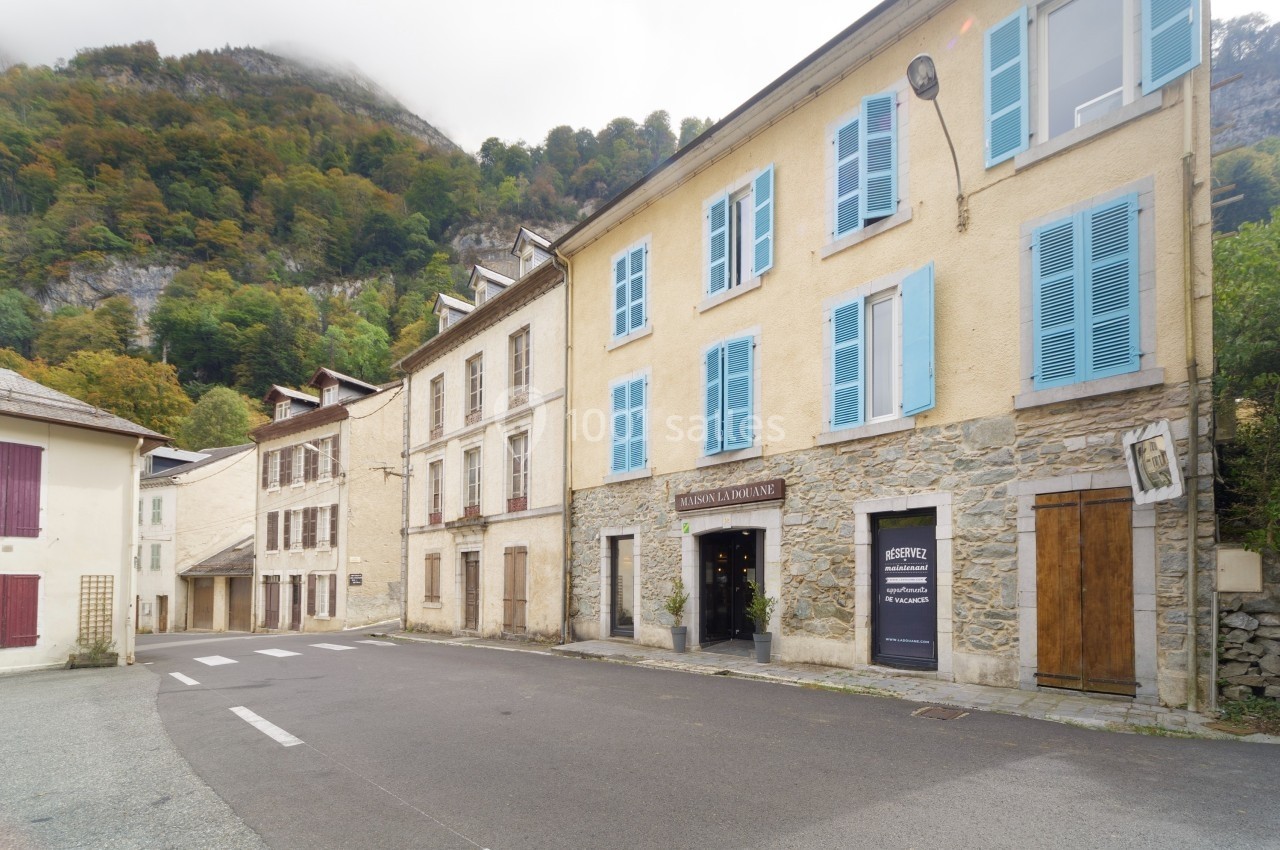 Rue calme bordée de bâtiments anciens aux volets colorés, avec une montagne boisée en arrière-plan.