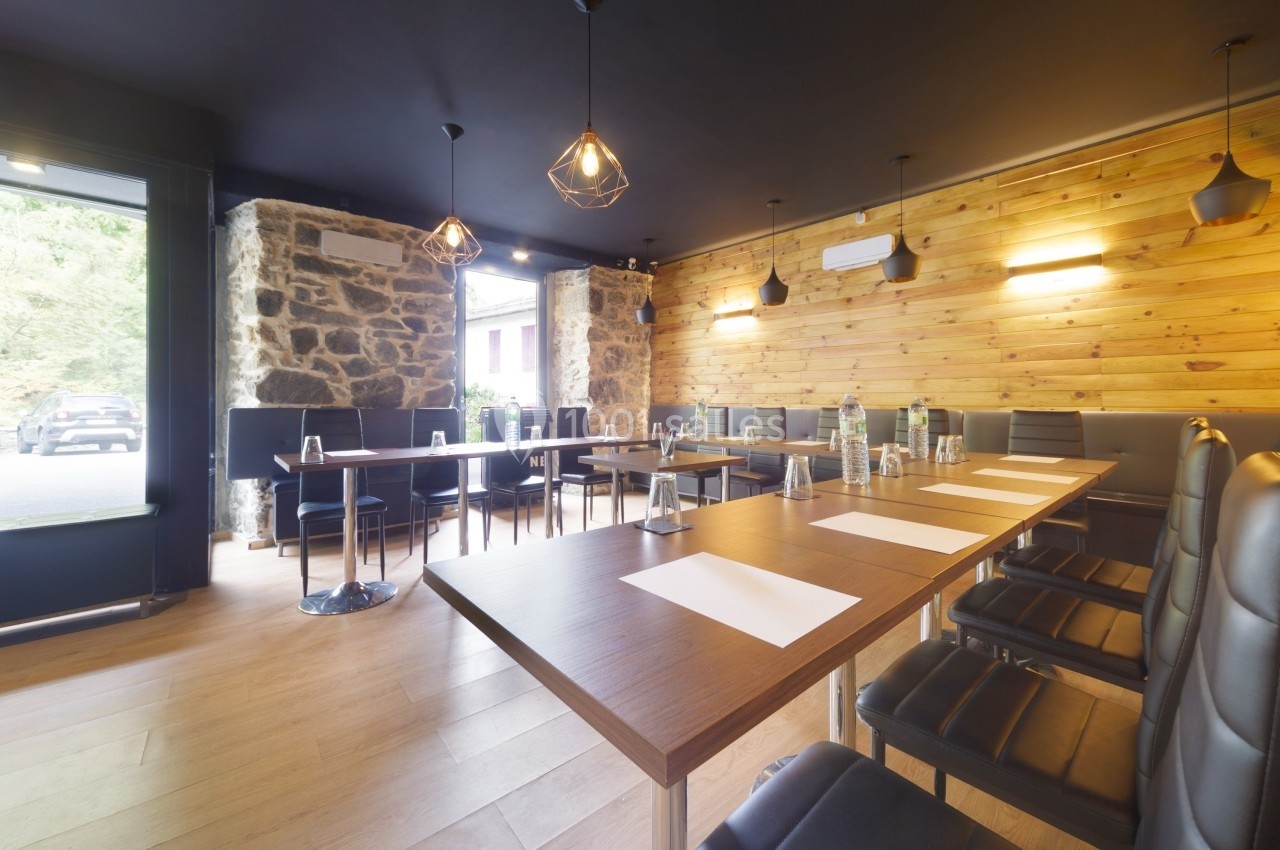 Salle de réunion lumineuse avec tables en bois, chaises noires, murs en pierre et bois, éclairage suspendu moderne.
