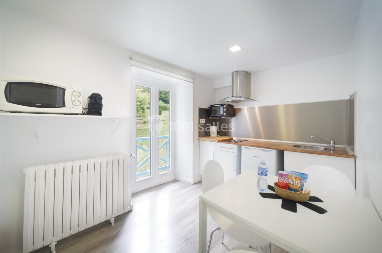 Cuisine lumineuse avec micro-ondes, cafetière, évier, table et chaises, donnant sur une fenêtre avec vue extérieure.