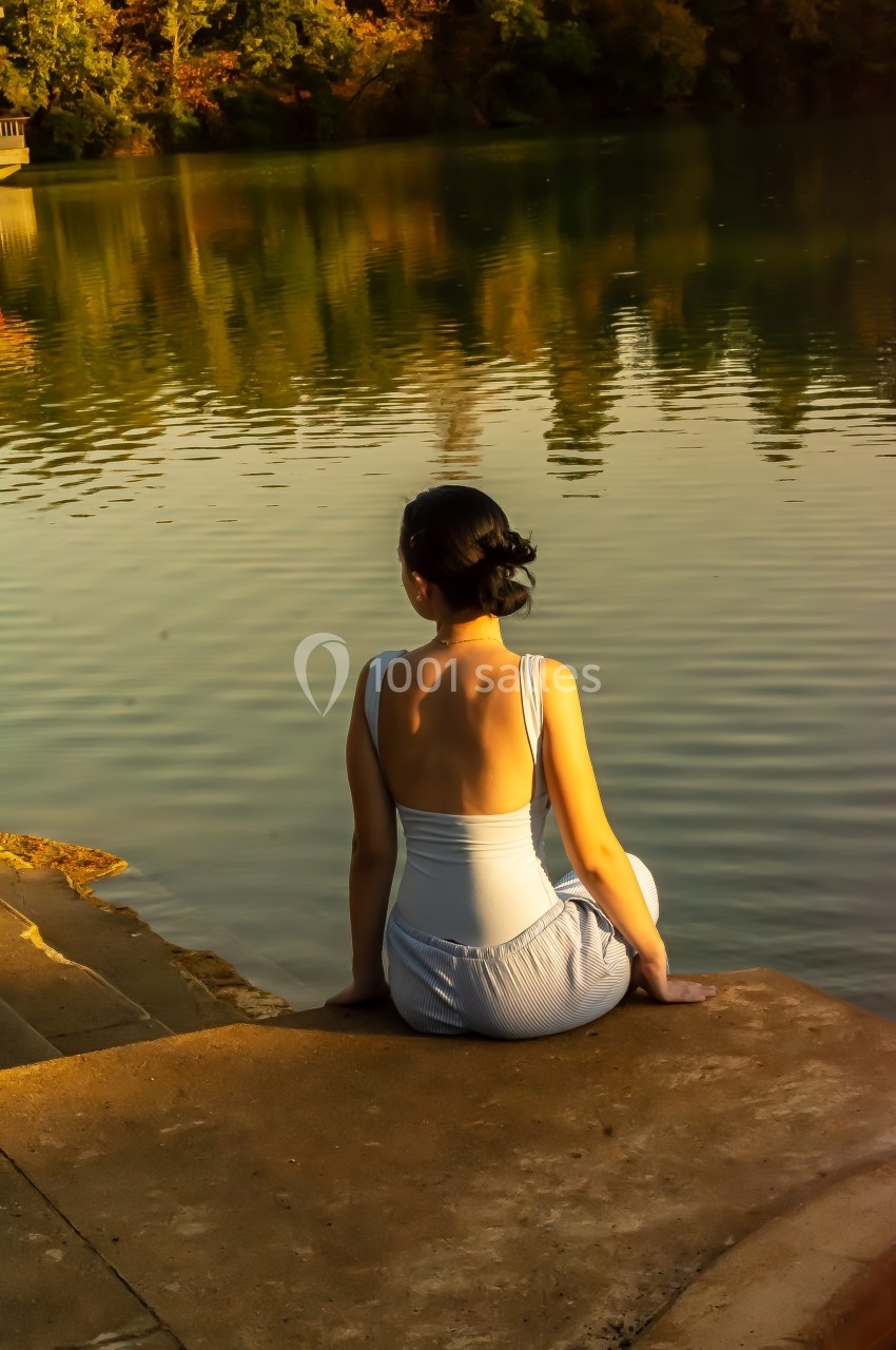 Femme assise au bord d'un lac, regardant l'eau calme entourée d'arbres sous une lumière dorée.