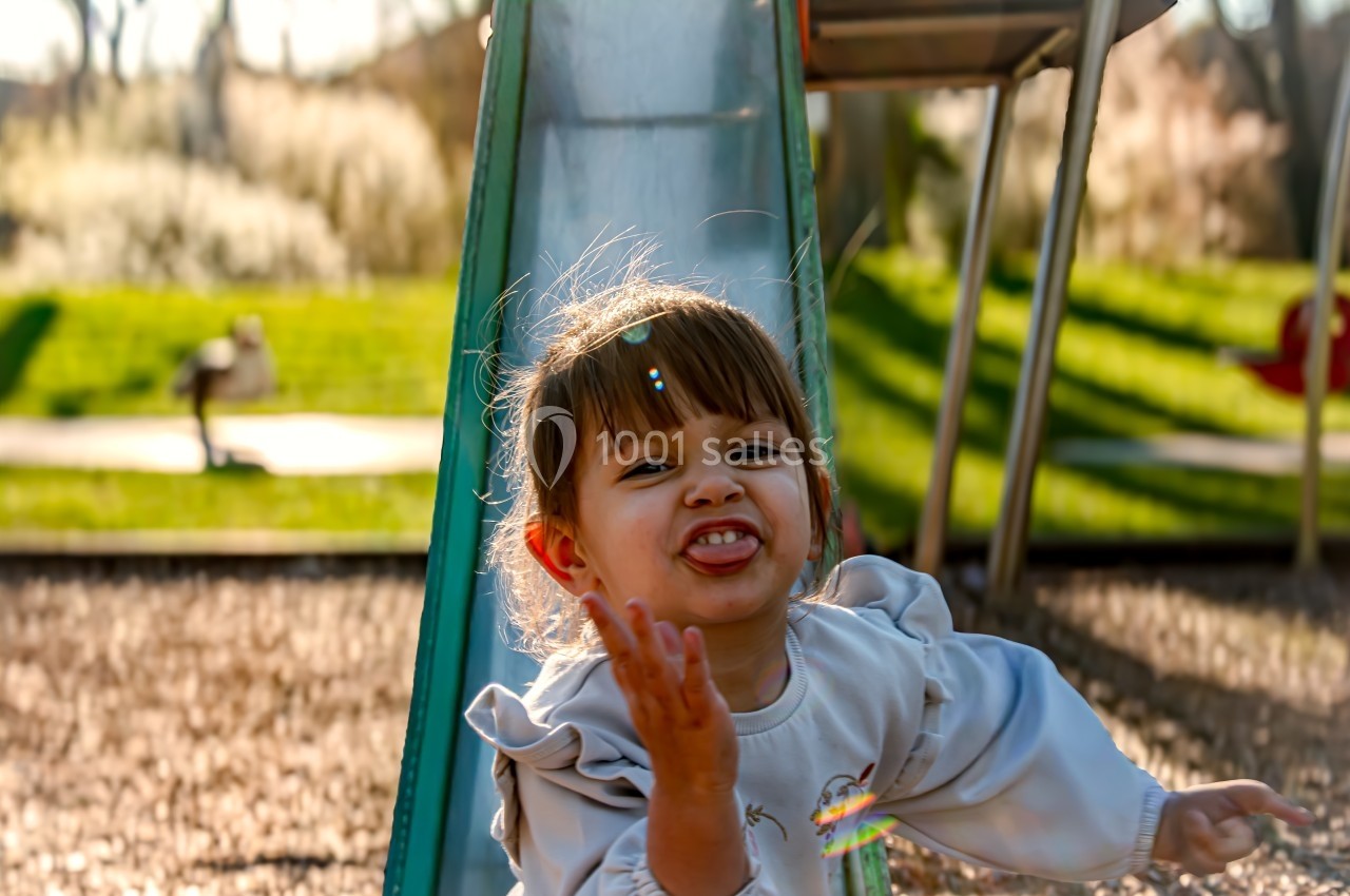 Une petite fille souriante joue devant un toboggan dans un parc ensoleillé.