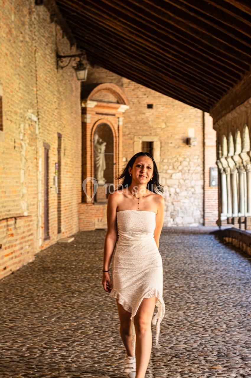 Une femme en robe blanche marche dans une galerie couverte en pierre avec des arches et des murs en briques.