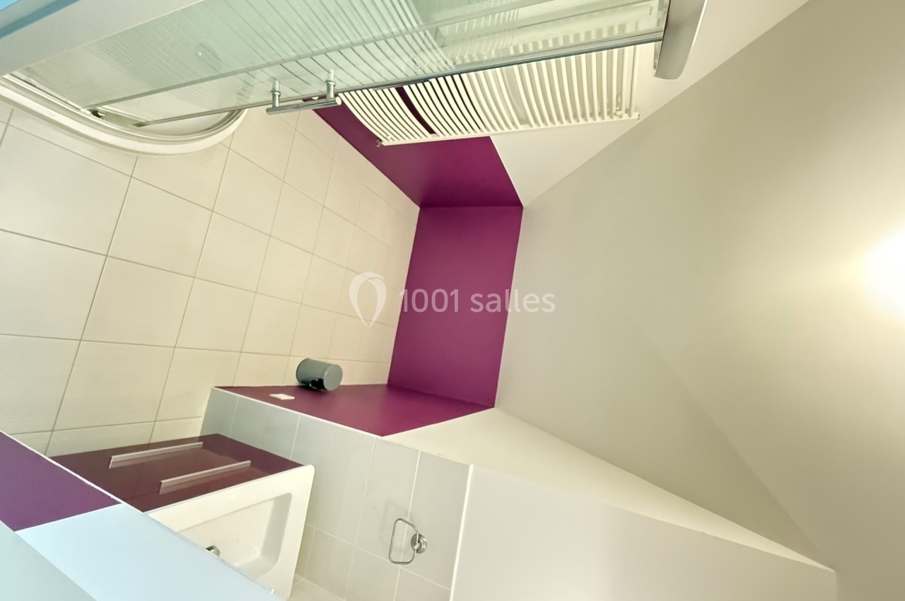 Salle de bain moderne avec murs blancs et violets, lavabo, miroir, radiateur et douche avec paroi vitrée.