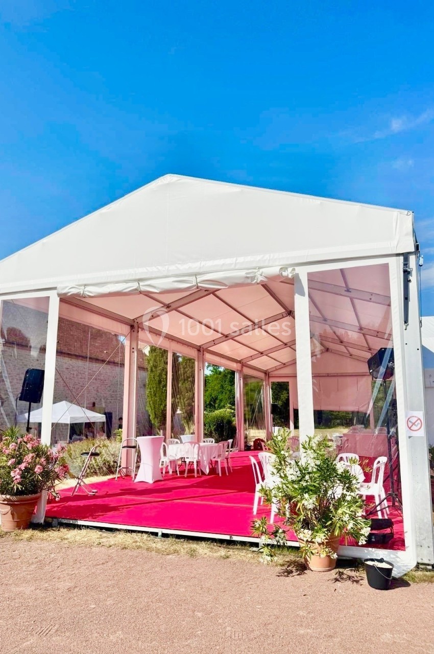 Tente blanche avec parois transparentes, sol recouvert de moquette rouge, tables et chaises blanches à l'intérieur.