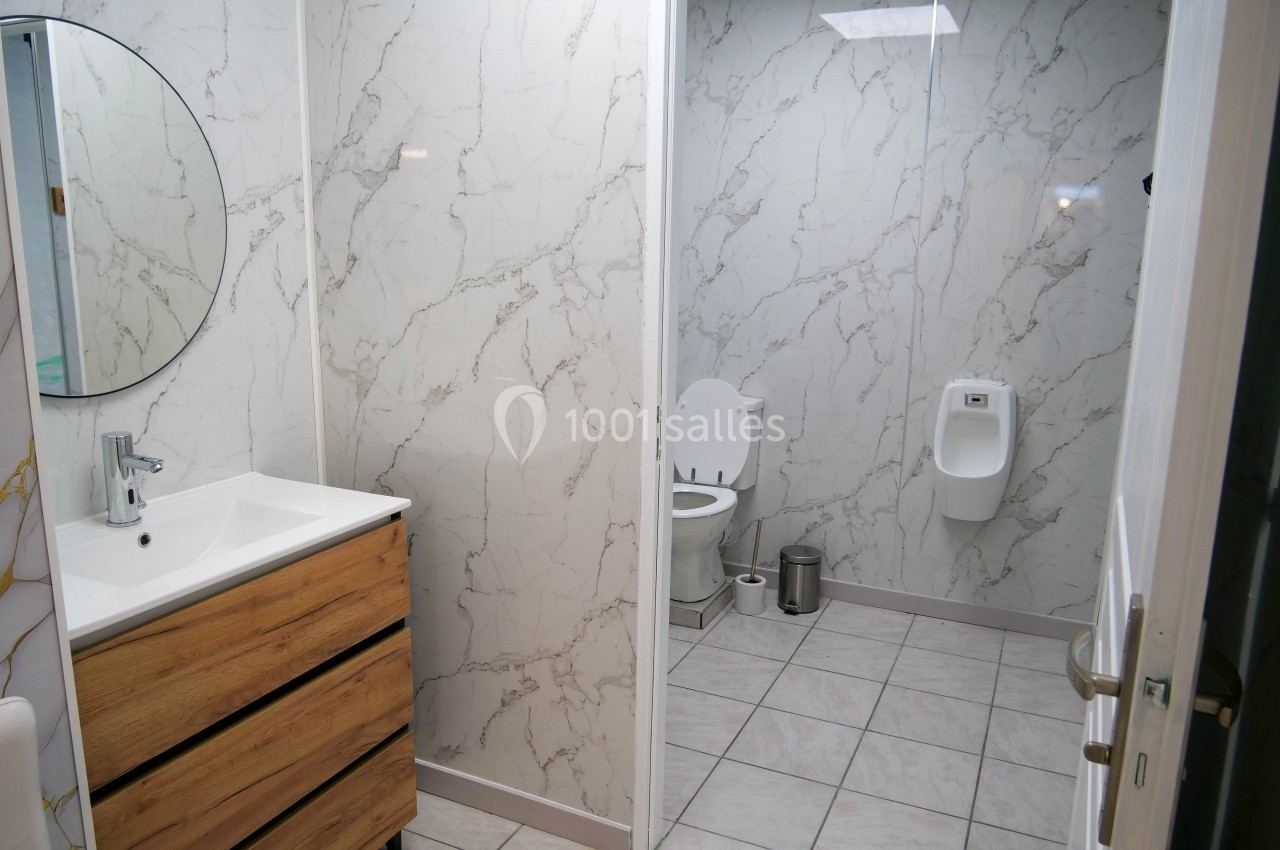 Salle de bain avec lavabo, meuble en bois, miroir rond, toilettes et urinoir, murs en carrelage effet marbre.