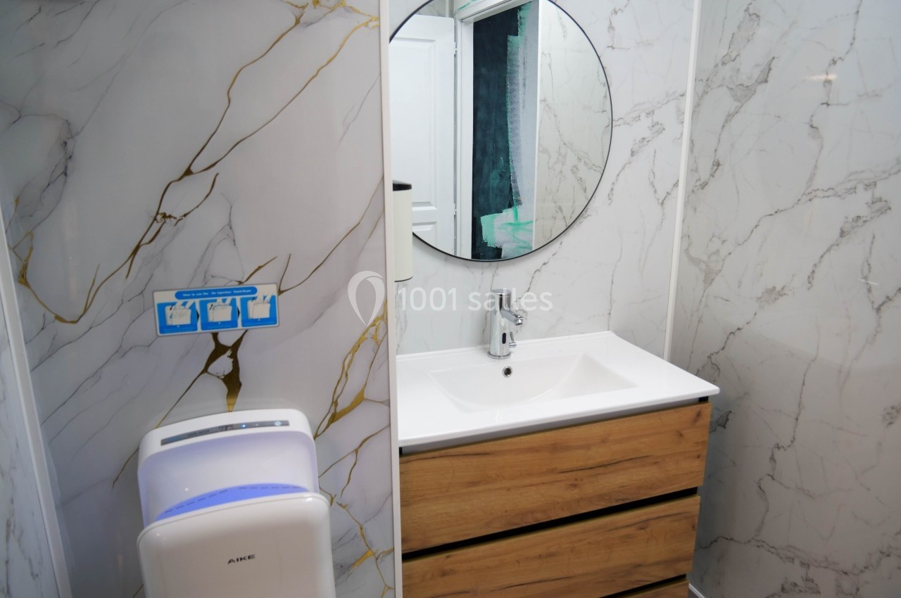 Lavabo avec meuble en bois, miroir rond et mur en marbre clair, sèche-mains mural à proximité.
