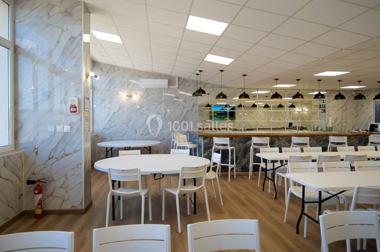 Salle lumineuse avec tables et chaises blanches, sol en bois clair, murs effet marbre et comptoir avec suspensions noires.