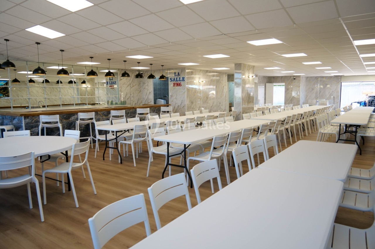 Salle à manger spacieuse avec des tables blanches alignées et des chaises, éclairée par des lampes suspendues.