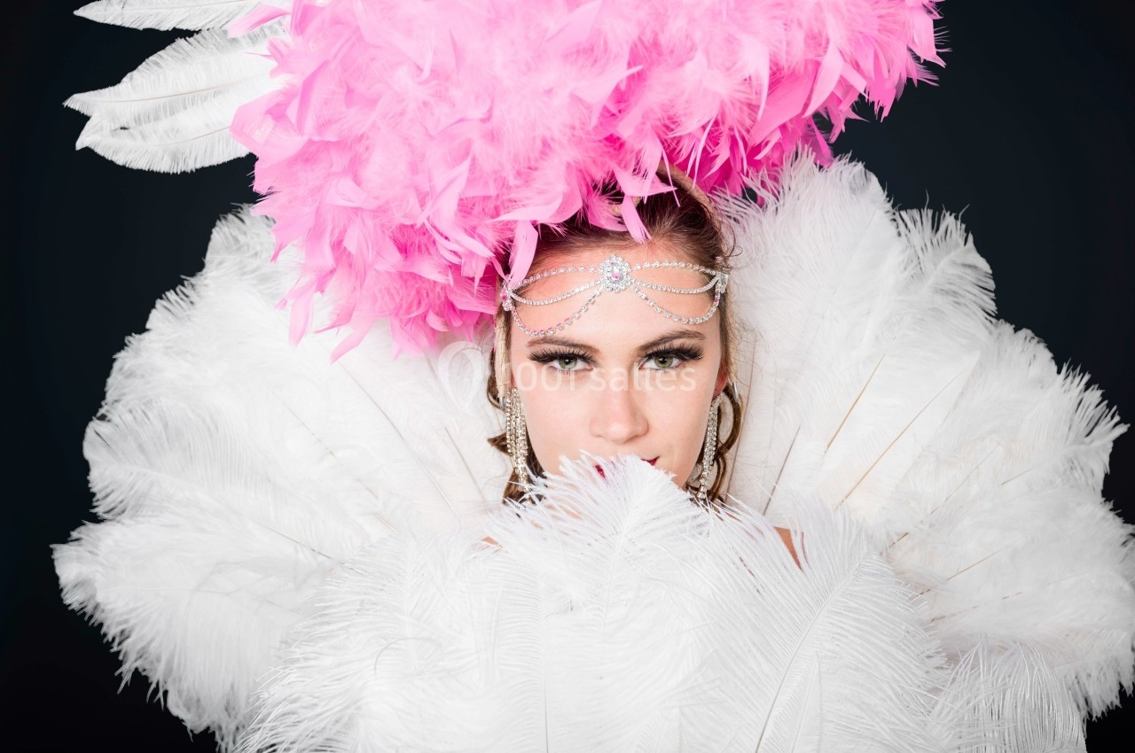 Femme portant une coiffe rose à plumes et un accessoire de tête argenté, entourée de plumes blanches.