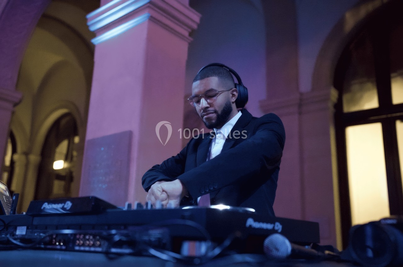 Un DJ en costume mixe de la musique sur une table de mixage dans un lieu avec des arches éclairées.