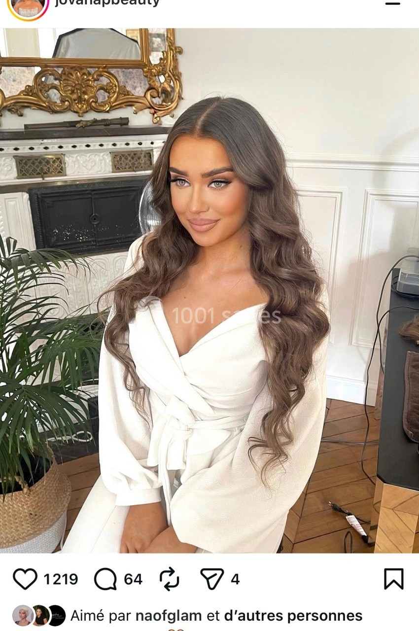 Femme assise dans un salon, portant une robe blanche, avec des cheveux longs et ondulés coiffés soigneusement.