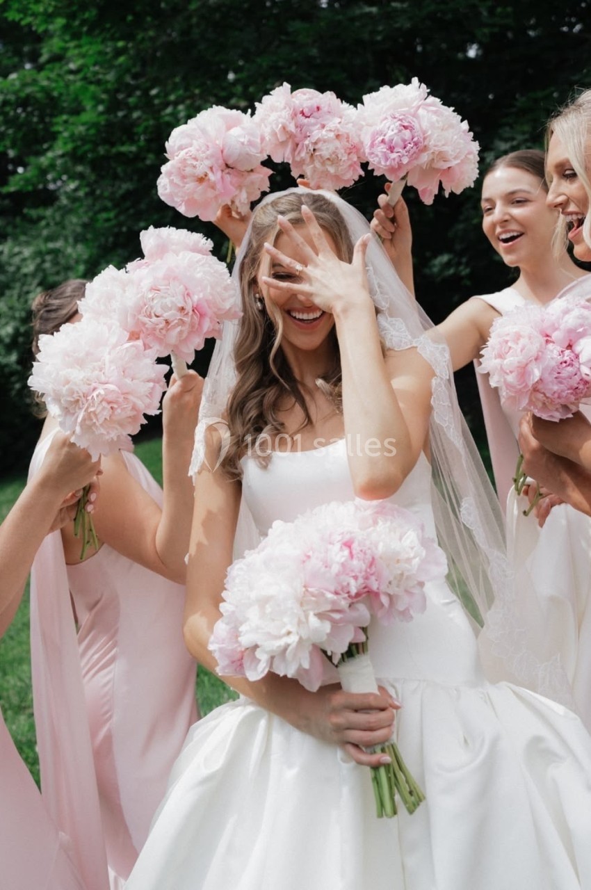 Une mariée souriante en robe blanche entourée de demoiselles d'honneur tenant des bouquets de pivoines roses.