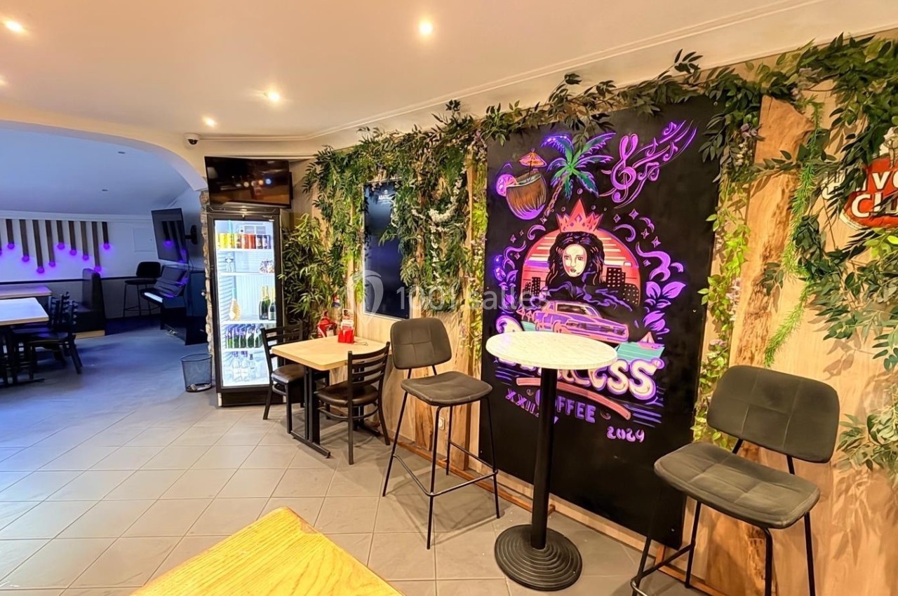 Intérieur d'un café avec décoration végétale, tableau mural coloré, tables et chaises modernes, et réfrigérateur en arrière…