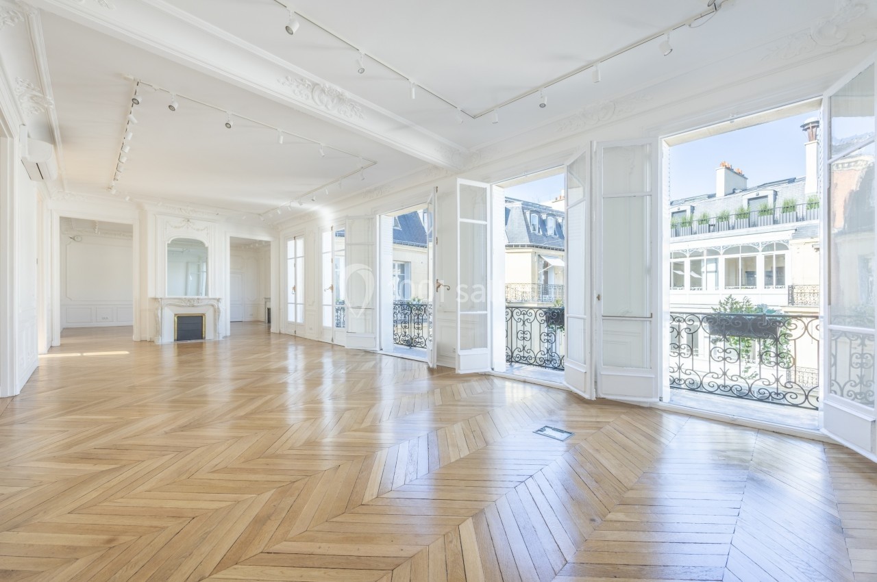 Salle lumineuse avec parquet en chevrons, grandes fenêtres ouvertes sur des balcons et vue sur des bâtiments voisins.
