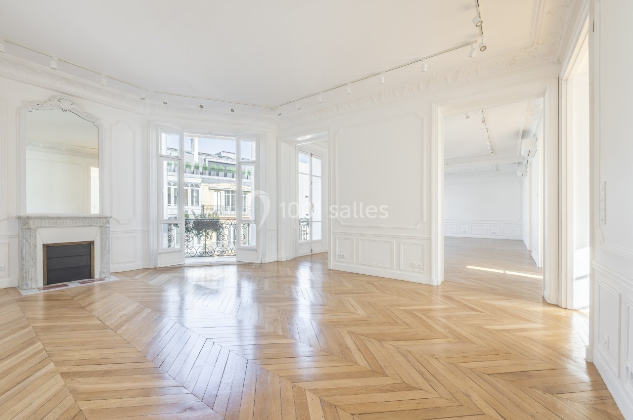Pièce lumineuse avec parquet en bois clair, cheminée décorative, moulures et grandes fenêtres donnant sur un balcon.
