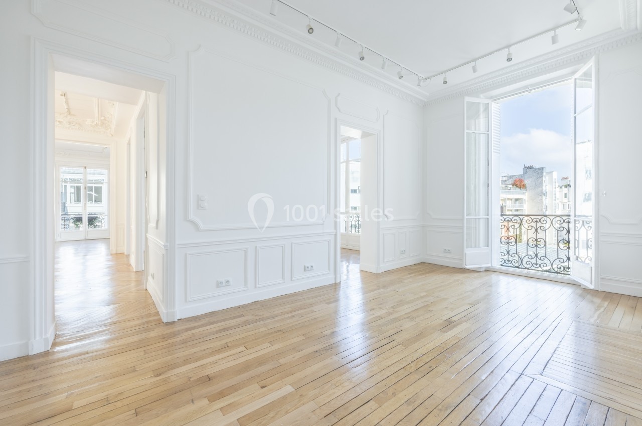 Intérieur lumineux avec parquet en bois clair, murs blancs, moulures, grandes fenêtres et balcon donnant sur la ville.
