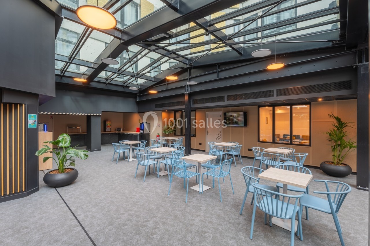 Espace lumineux avec tables et chaises bleues sous une verrière, entouré de plantes et d'un comptoir au fond.
