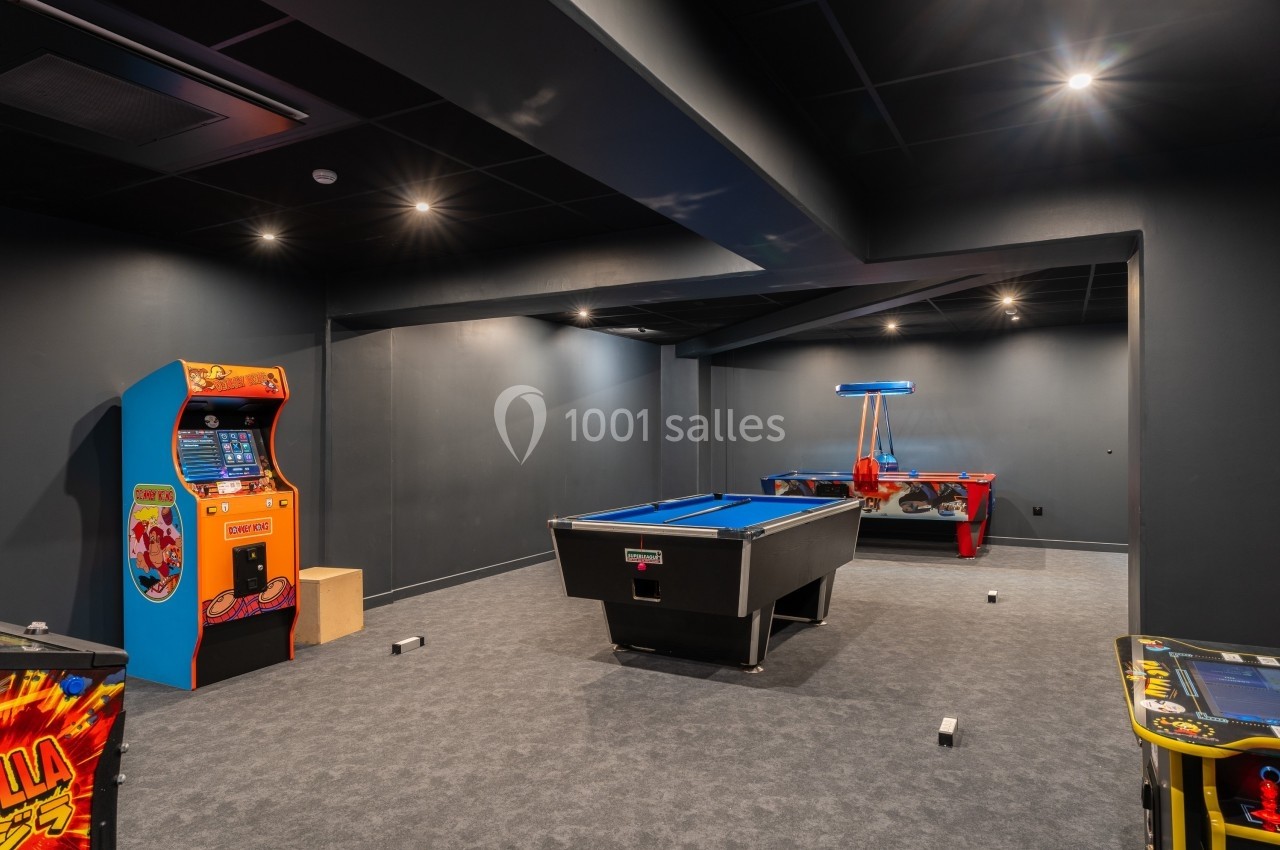 Salle de jeux avec un billard, une table d'air hockey et des bornes d'arcade dans un espace sombre et moderne.