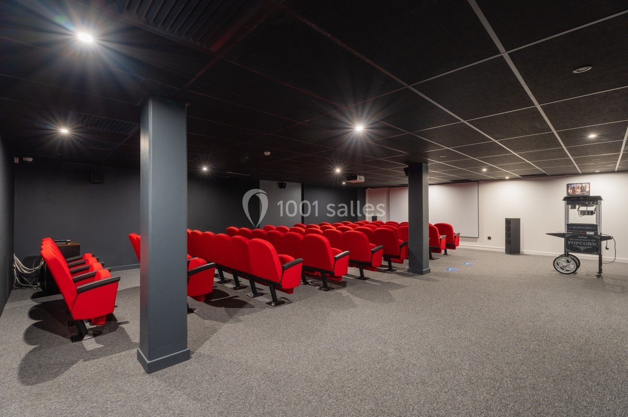 Salle de projection avec rangées de sièges rouges, écran blanc à l'arrière et équipement audiovisuel sur la droite.